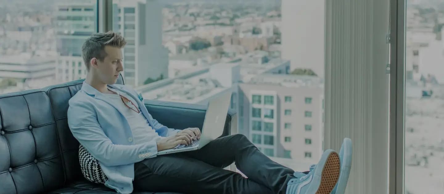 Person wearing a light blue blazer and black pants, sitting on a black couch, working on a laptop with a cityscape visible through the window in the background.