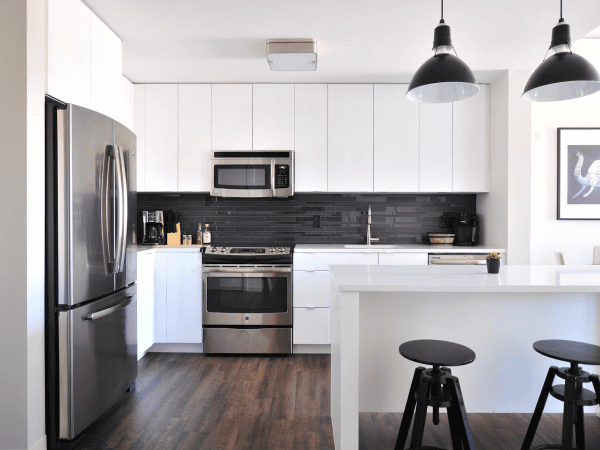 A modern kitchen with white, flat-front cabinets and stainless steel appliances. Light wood floors and a breakfast bar with two stools complete the look.